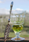 A reservoir glass filled with a naturally colored verte next to an absinthe spoon.