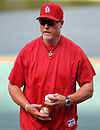 A man with a whitening goatee wearing a red baseball cap and short-sleeved warm-up shirt, holding a baseball in front on him in each hand