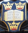 A shield displaying a coat of arms; on a dark blue background, an open book displays the words "Dominus Illuminatio Mea"; two gold crowns above, one below