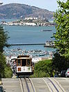 Front-end photograph of one of the San Francisco Cable Cars cresting a hill. A panorama of San Francisco Bay in the background features Alcatraz Island, another National Historic Landmark.