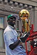 A black man with sunglasses on is holding a trophy at a parade