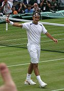 A dark-haired man is waving to the crowd with his tennis racket in his right hand, and he is wearing all white clothing
