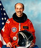 A Caucasian male astronaut in front of the American flag wearing an orange jumpsuit while holding a helmet.