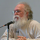 A Caucasian male with gray hair and a white beard and mustache sitting in front of a microphone while wearing a white long sleeved shirt and glasses.