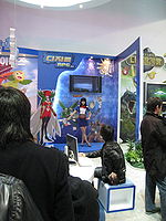 Inside a large, brightly lit convention center room with white walls is positioned a promotional display booth for a video game. A saleswoman clad in a blue shirt and skirt and a red bowtie motions towards several illustrations on the booth, explaining their implications. The illustrations are anime-styled and depict several outlandish and brightly colored creatures. Three men in dark jackets, one at a, watch the demonstration.