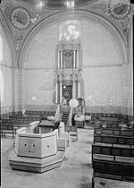 A sepia photograph shows the interior of the synagogue. In the foreground lies the reading desk atop a simply paneled almemar. Rows of wooden benches line the right and left side of the nave. The holy ark, set in the centre of the eastern wall, is decorated with baroque carvings and set off against four Corinthian columns. Its top reaches a large clover-shaped window, which sits just below one of the four supporting arches. The walls faintly show decorative murals, with two large round frescos situated at the top left and right corners.