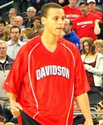 A young man in his early 20s is warming up for a basketball game. He is wearing a red warm-up jersey with "Davidson" written in white across the chest. He is a light-skinned male of mixed race (half white by his mother, half African American by his father).