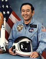 An image of LTC Onizuka with a model of the Challenger shuttle and astronaut helmet on a desk in front of him. The United States Flag, and a smoky blue backdrop in the background.