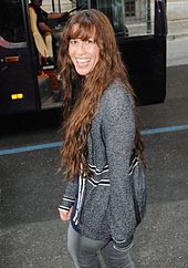 The picture of a woman who is smiling. She is walking on a street and she is looking back over her left shoulder. She wears a grey sweater with some black frames, and jeans of the same colour. In the background a black bus is visible.