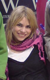 A woman in late 20s with blonde hair and brown eyes, smiling, wearing a white T-shirt, a black tank top and a pink scarf.