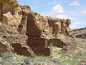 Cliff-side set of ruined walls in daytime. In front of a cliff running diagonally from near left to middle right, rectangular slabs of stone, each somewhat smaller than a common brick, are stacked to compose a wall. Walls are seen delimiting several smallish rectangular "rooms". In the background at middle-right, a set of stone steps is seen leading up from the walls to the top of the cliff.