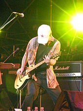 Close-up of a man onstage with a guitar wearing jeans and a baggy shirt. His head is lowered and his face is obscured by a white hat with a red star on it. In the background is musical equipment.