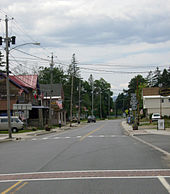 A two-lane highway passes alongside several two-story homes, some of which house businesses. One side of the highway is lined with telephone poles that have streetlights and American flags mounted on them.
