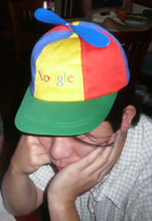 Asian man in his twenties wearing a blue, green, yellow and red propellor hat that says "Noogle"
