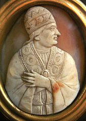 Cameo of the upper body of an older jowled man looking to the side. He is wearing papal vestments and a conical skull cap, while his left hand is holding the robe closed.