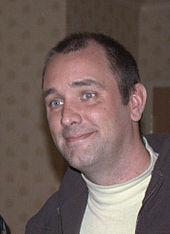 A brown-haired man wearing a lime shirt and grown jacket smiles and looks at something off-screen.