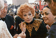 An aged Ball standing in a crowd of celebrities, wearing a black and gold sequinned dress with her characteristic red hair, looking fragile.