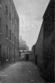 An old, tall, brick wall on the right of a courtyard, and a four-story, dismal-looking, brick building on the left. A young boy is standing in one of the doorways of the building.