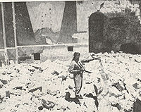 A sepia photograph shows a soldier, gun in hand, standing amidst the rubble of the destroyed synagogue. Behind him, remnants of the eastern wall shows a painted fresco of Mount Sinai and two arched tablets symbolising the ten commandments.