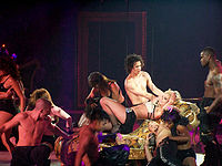 Image of a blond woman. She is laying in a flowered divan, with her body extended and her hands in her head. She is wearing a sparkly bra and lingerie, with black high-heeled shoes. The divan is attached to wires. She is surrounded by several people, including bare chested men of differentes races. Several women also surround her, wearing black lingerie ensembles. Below her, an Hispanic man and an African American woman are touching each other in a sexual way.