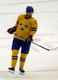 An ice hockey player skating relaxed on the ice. He leans slightly backwards with one skate lifted off the ice and is looking downwards to the left. He wears a yellow jersey with blue trim and a blue, visored helmet.