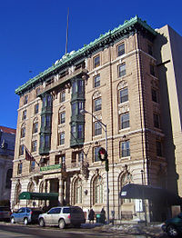 An ornate tan-colored brick building six stories tall seen from across a city street and to its right. There are cars and some piles of old snow in front of the building.