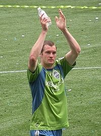 the head and torso of a young man, wearing a green and blue top and blue shorts, standing on a grass field. He has his arms raised above his head in what appears to be a clapping motion.