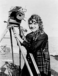 Picture of a very pretty young lady with long blonde ringlets. She is smiling and wearing a Scottish outfit while standing next to a motion picture camera near the ocean.