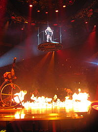 Image of a stage. In the center, a blond woman wearing a leotard and a black hat is in mid-air, standing inside a circular platform suspended by wires. The center of the stage is covered with fire. Several performers are in the sides of the stage, doing acrobatics in jungle gyms.