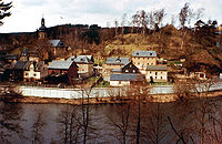 View of a small village with houses and a church, located in a wooded valley, with a river in the foreground. A high concrete wall separates the village from the river.