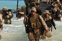 color photo of two columns of Marines wade through waist deep water disembarking from a landing craft onto a beach