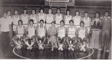 A black and white photograph of people standing in two rows in front of a basketball hoop. The back row mostly has men wearing white sleeveless shirts and white shorts while standing, while the front row has men wearing white sleeveless shirts and white shorts seated on chairs except for a woman in the center wearing a dress.