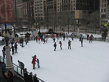 Part of an ice skating rink viewed from above with dozens of skaters to one side mostly standing behind cones on the ice.