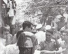 A crowded and narrow street with a lot of people in the vicinity. A tall man with brown hair and sunglasses stands in front of a smaller Asian man with black hair, shielding a shorter man in light shirt with a bloodied face. The smaller Asian man puts an open palm in the air to acknowledge the tall man. A steel-helmeted policeman is in the foreground and others in white caps are in the background.