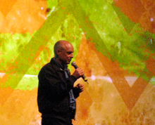 A bald man speaking into a microphone is standing in front of an abstract painting containing blotches of orange and lime green and corrugated lines.