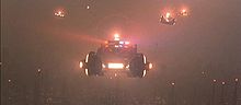 A screenshot from the film shows a line of police vehicles with flashing lights flying high above a smog-covered cityscape. Below them several small pinpoints of light from aircraft-avoidance lights on the tops of towers are all that can be seen of the city