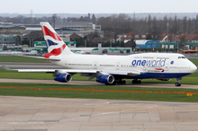 A British Airways Boeing 747-400 in Oneworld livery taxiing on the taxiway, with Heathrow Airport facilities in the background and green grass patch in the foreground