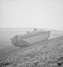 A tracked armoured vehicle drives up the bank of a river.  In the background lies the wrecked frame of a metal bridge.