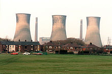 Three massive grey-taupe coloured funnels dominate the scene in which there is a flat piece of green grass in the foreground, red-brick housing in the midground, and behind them in the background the industrial funnels against a white sky. The funnels appear three or four times the size of the row of terraced properties in the midground, and stand independently of each other. Associated with them are two chimneys standing at similar height and a control building in the hazey distance.