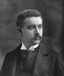 A man with dark hair and a mustache wearing a dark jacket and vest, patterned tie, and white shirt