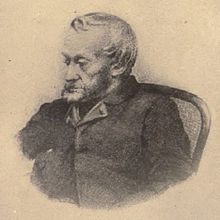 Black-and-white depiction from the waist up of an old, bearded man dressed in a dark jacket, sat in a chair and looking towards the left.