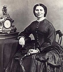19th century photograph of a woman seated with her arm resting on a table. Her dark hair is neatly partedin the middle.  She is smiling slightly