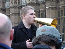 A picture of Dan Bull in Parliament Square, holding a handheld megaphone, standing on a raised platform and addressing protesters