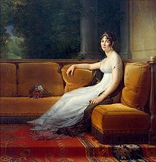 Woman with brown hair, in a white dress and tiara, sitting on a plush orange sofa