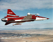 Starboard view of red and white single-engine jet fighter aircraft banking banking left. At the wingtips are missiles.