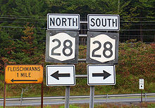 Two road signs with the number 28 in black on a white background with "North" and "South" written over them and arrows pointing in opposite directions underneath. On the left is a sign saying "Flesischmanns 1 mile" in black on a gold background with an arrow pointing to the left. Behind the signs are roads, woods and telephone wires
