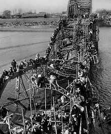 Black-and-white photo of people crossing a river via a destroyed brdige