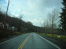 Ground-level view of a two-lane road with numerous homes atop a slight hill on the left-hand side. A field with a few trees is on the right.