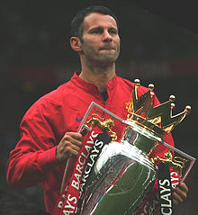 A head-and-torso photograph of a man with dark hair wearing a red tracksuit top. He is holding a large silver trophy with a gold crown on top. The trophy is decorated with one red ribbon and one black ribbon attached to each handle.