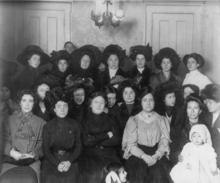 A large group of women wearing dark, early 20th century clothing pose for the camera. Two men peek in from behind the posed group, and one of the women holds a child.